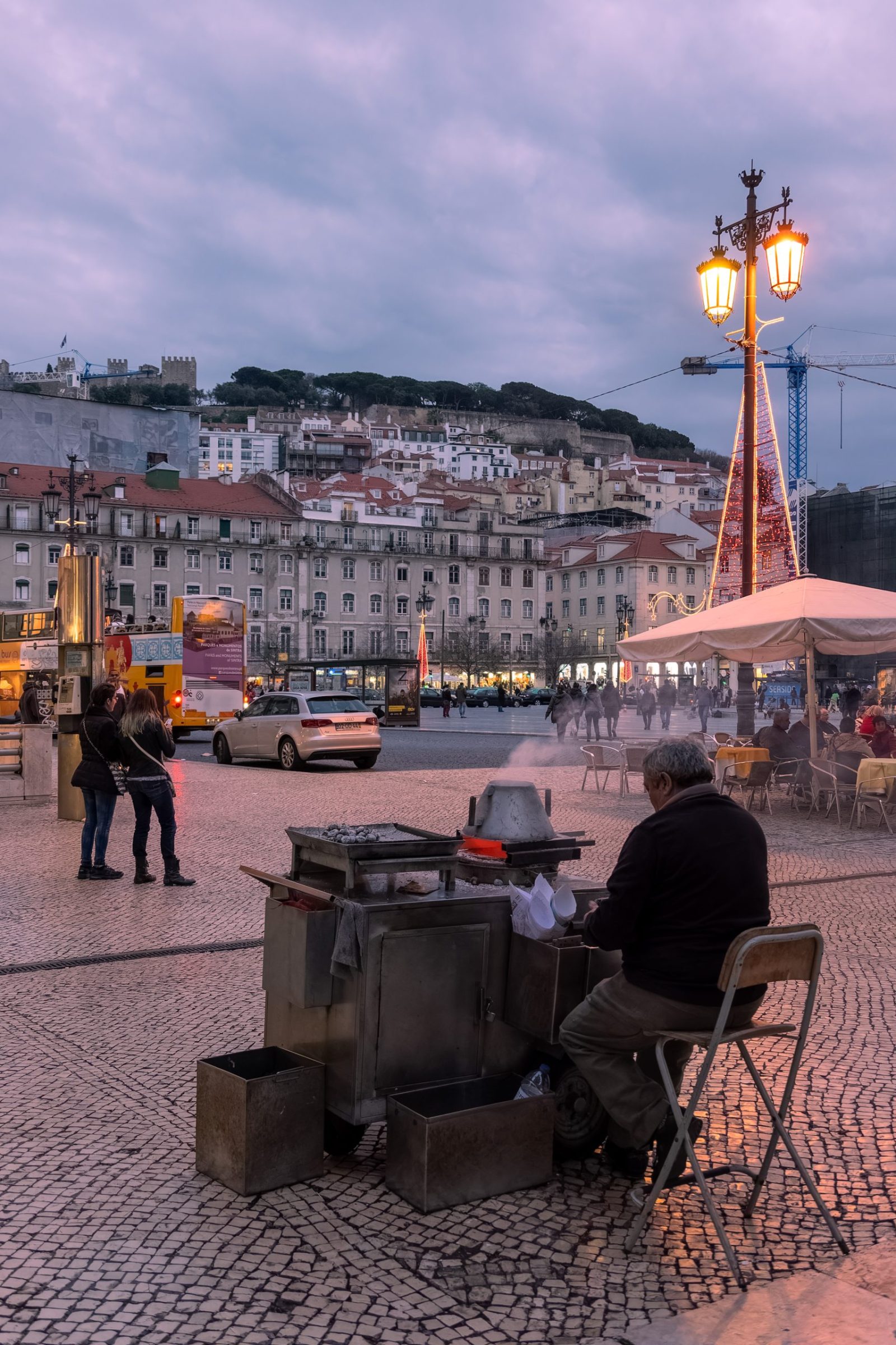 A roasted chestnut cart at dusk in Praça da Figueira with São Jorge Castle behind